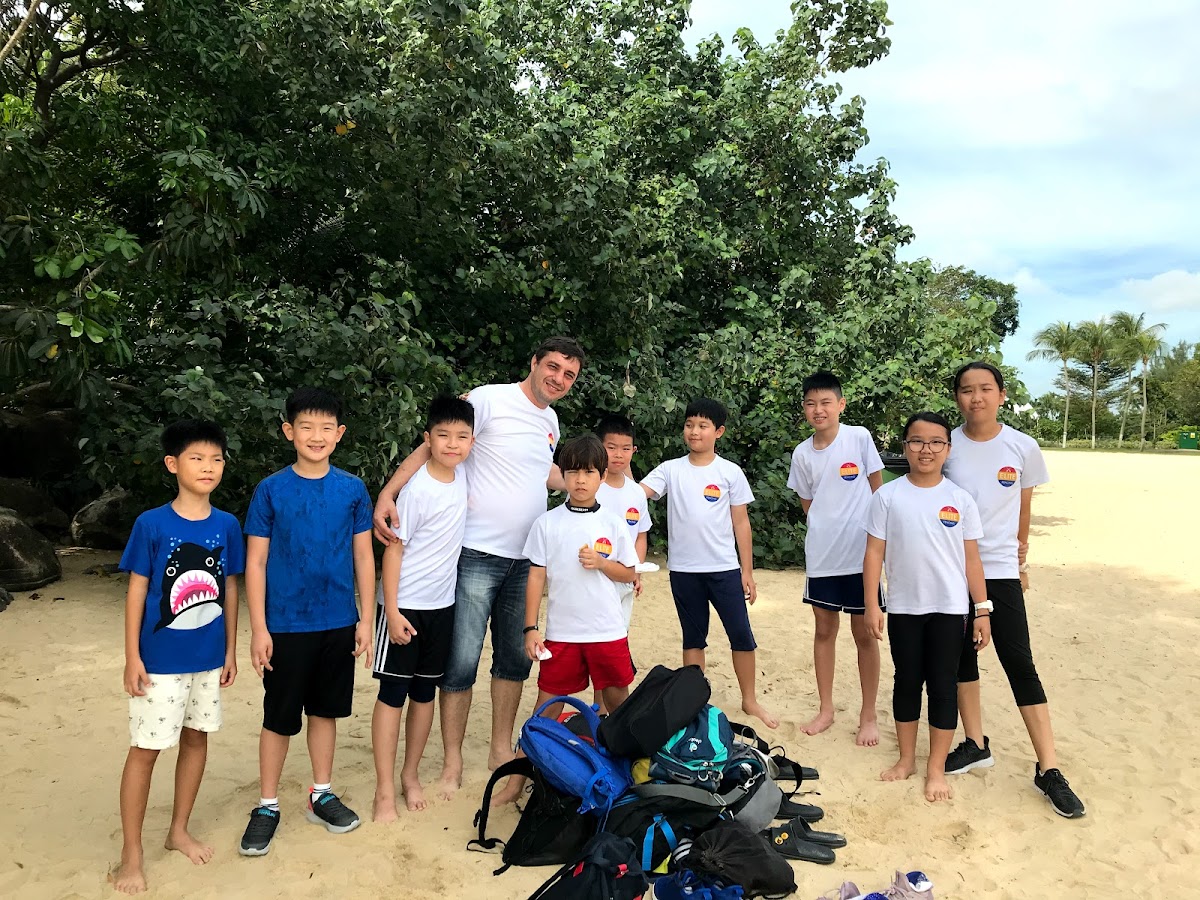 A group of children and a male trainer are posing for a photo outdoors on a sandy area with trees in the background.