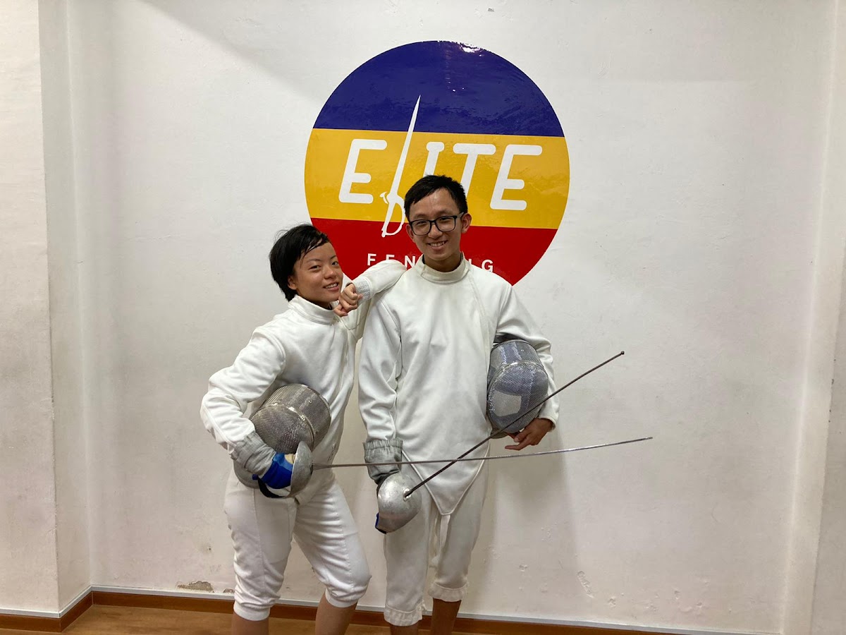 Two fencers in white uniforms pose with swords in front of a wall featuring the 'Elite Fencing' logo.