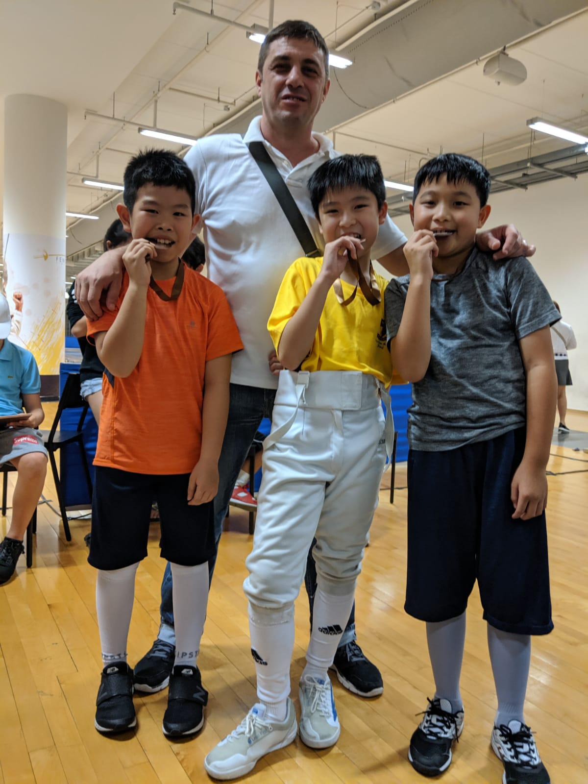 A male trainer poses with four young boys who appear to be practicing fencing in an indoor gym setting.