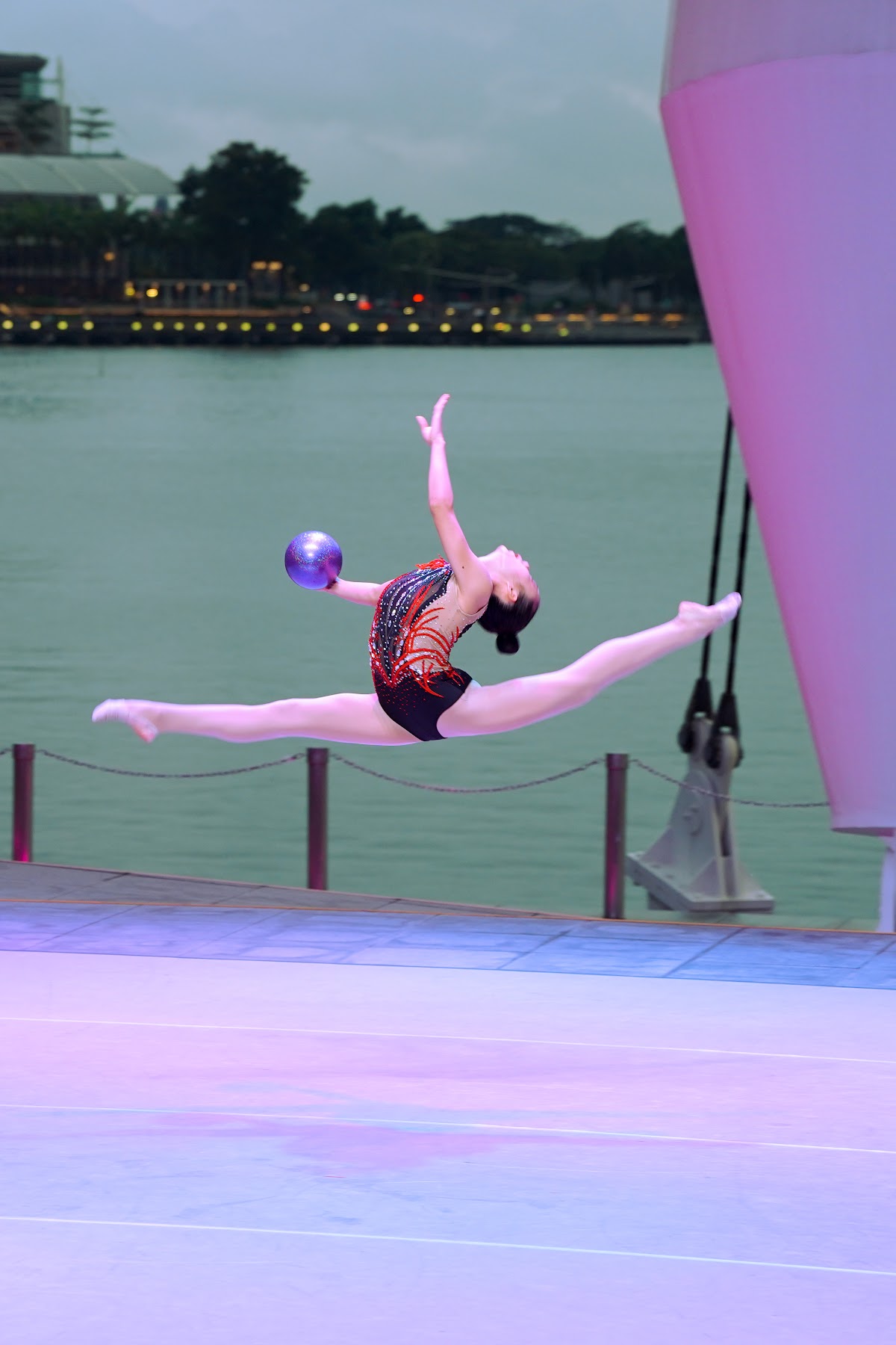 A female gymnast performs a dynamic routine with a ball on a stage near water.