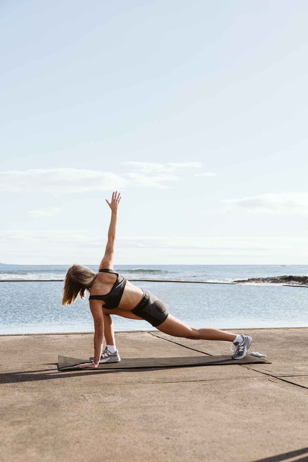 A woman is performing a plank exercise on a yoga mat outdoors near the ocean.
