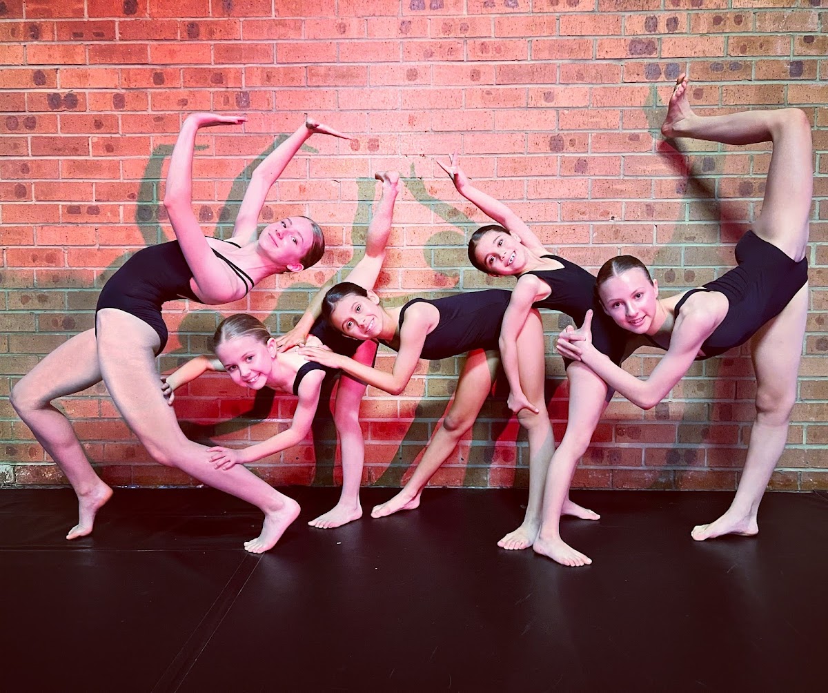 A group of young girls in leotards are posing and stretching against a brick wall during what appears to be a dance or gymnastics class.