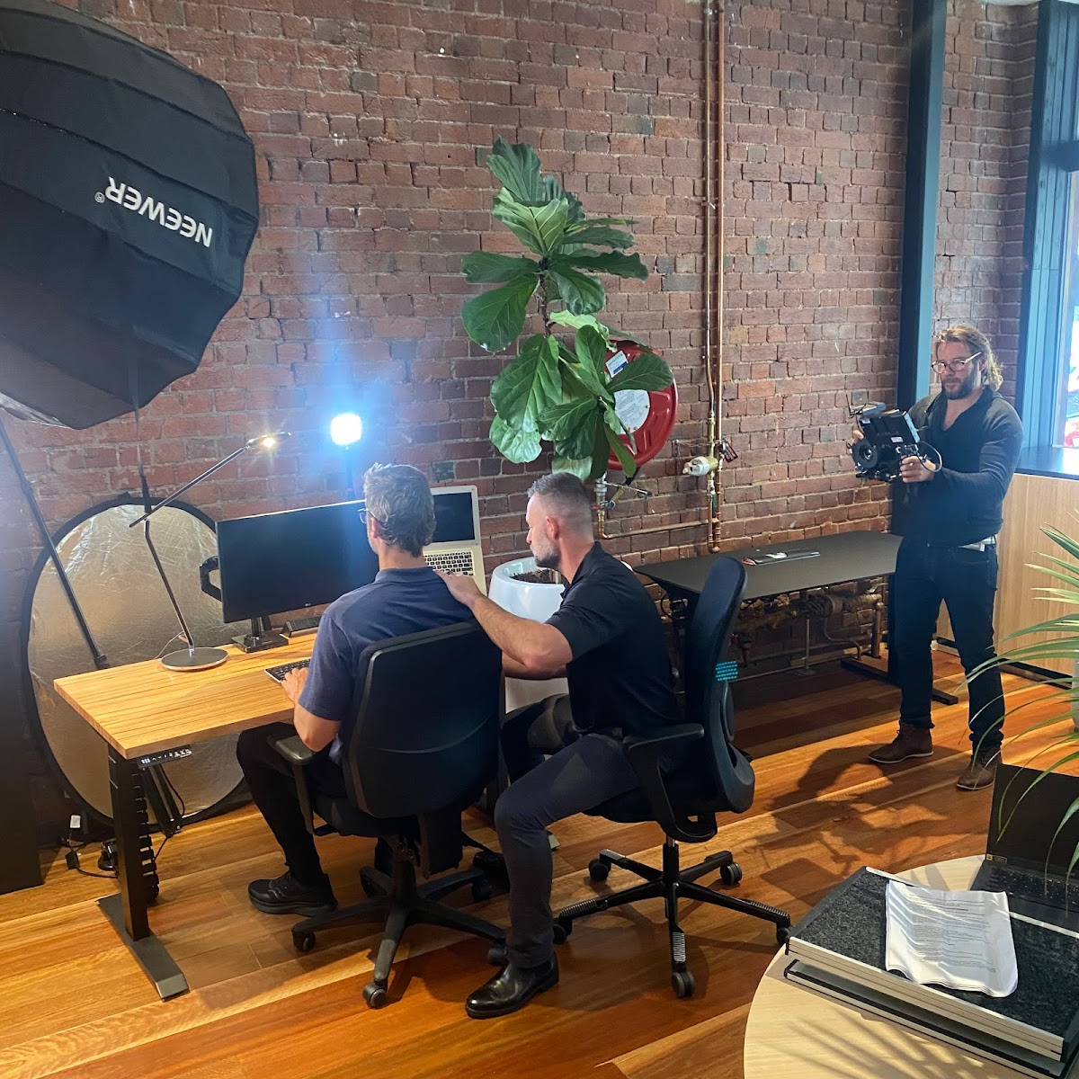 A studio setting with two men seated at a desk, seemingly in a meeting, while a third man films them with a camera and lighting equipment.