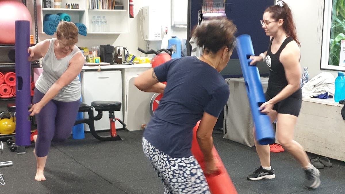 Three women are participating in a small group fitness class using yoga mats and free weights in a brightly lit, minimalist gym space.