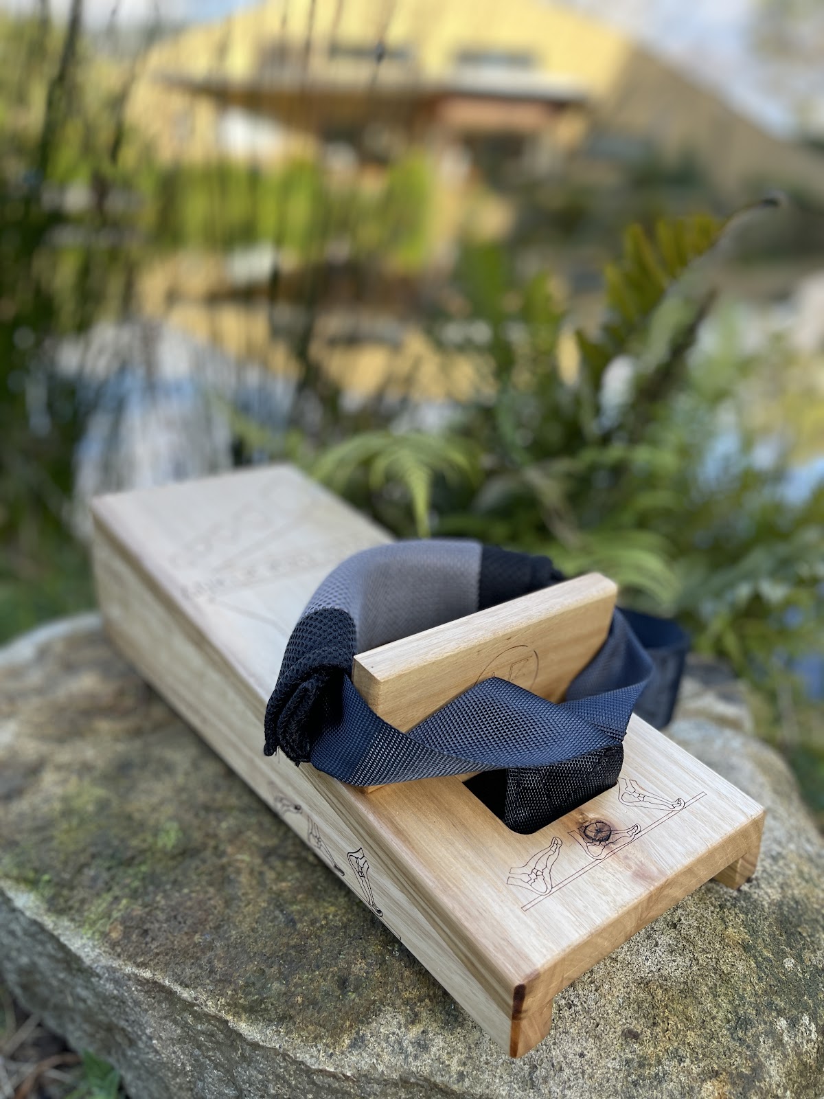 A wooden reformer pilates machine sits on a rock in an outdoor setting with greenery and water visible in the background.