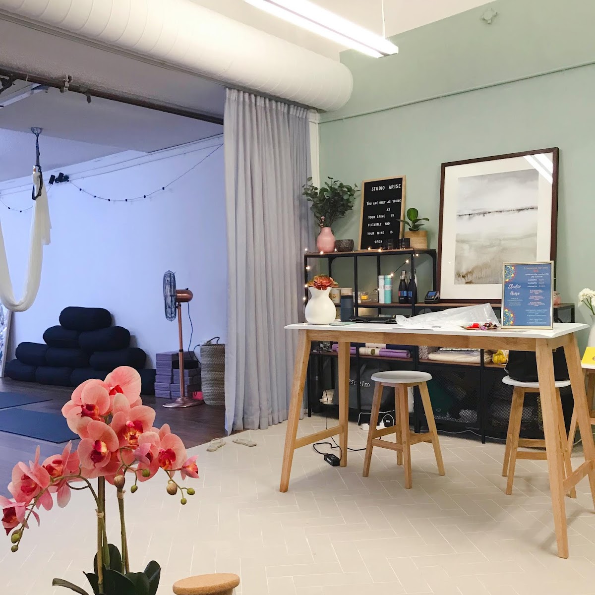 The photo shows a serene studio space with a reception area featuring a table, stools, and decorative plants, with a yoga area visible in the backgrou