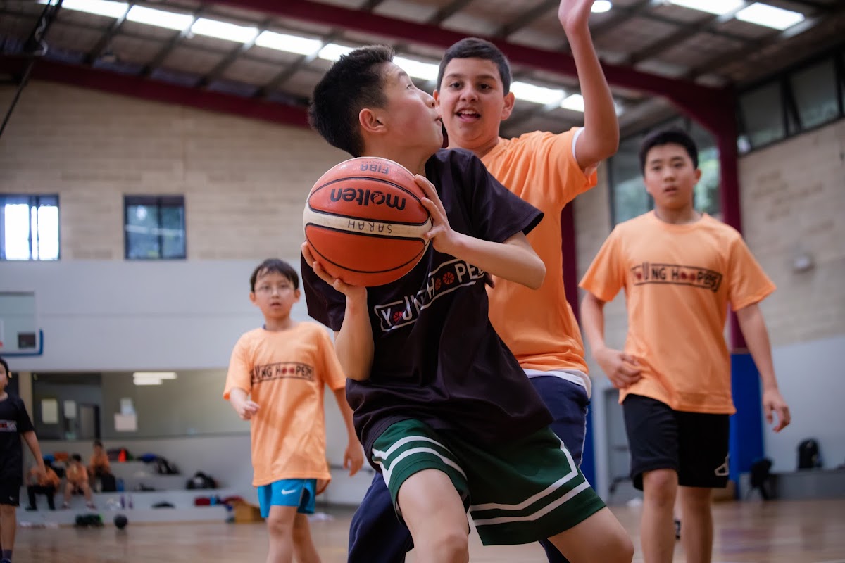 A group of young children are playing basketball in a brightly lit indoor gym.