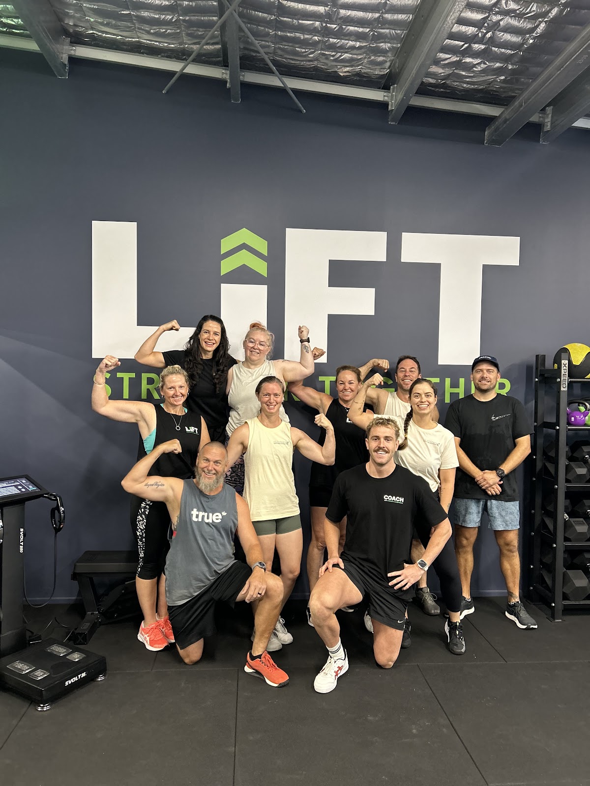 A group of gym members and trainers pose for a photo in a fitness studio with boxing bags visible, showcasing strength training.