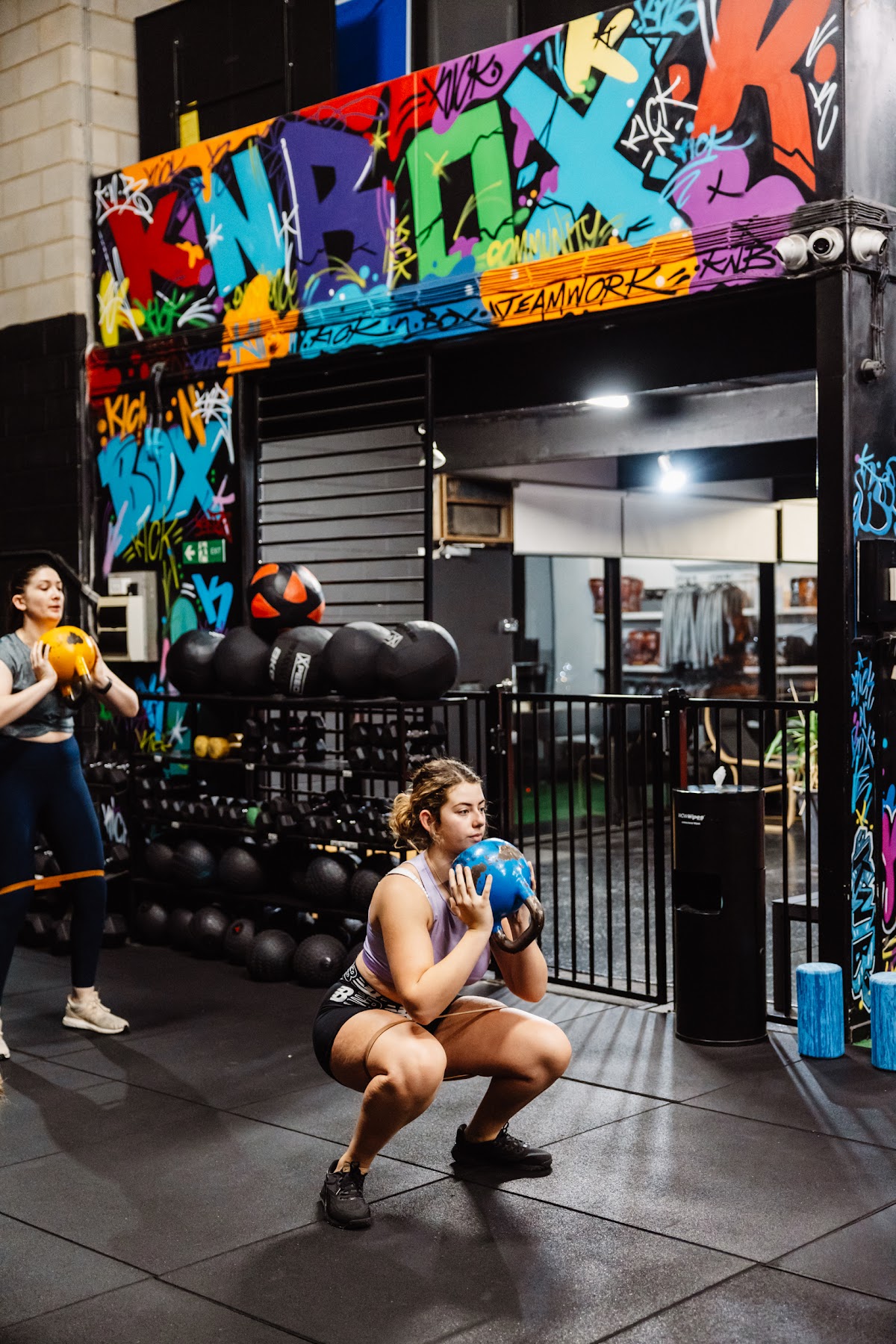 Two women are performing kettlebell squats in a gym with industrial decor and boxing bags visible.