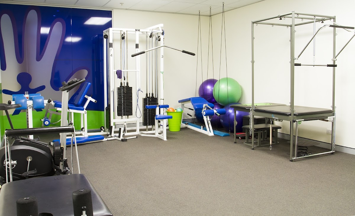 The photo shows a clean, minimalist gym interior with cable machines, free weights, and TRX suspension trainers, featuring a wall mural and a grey car