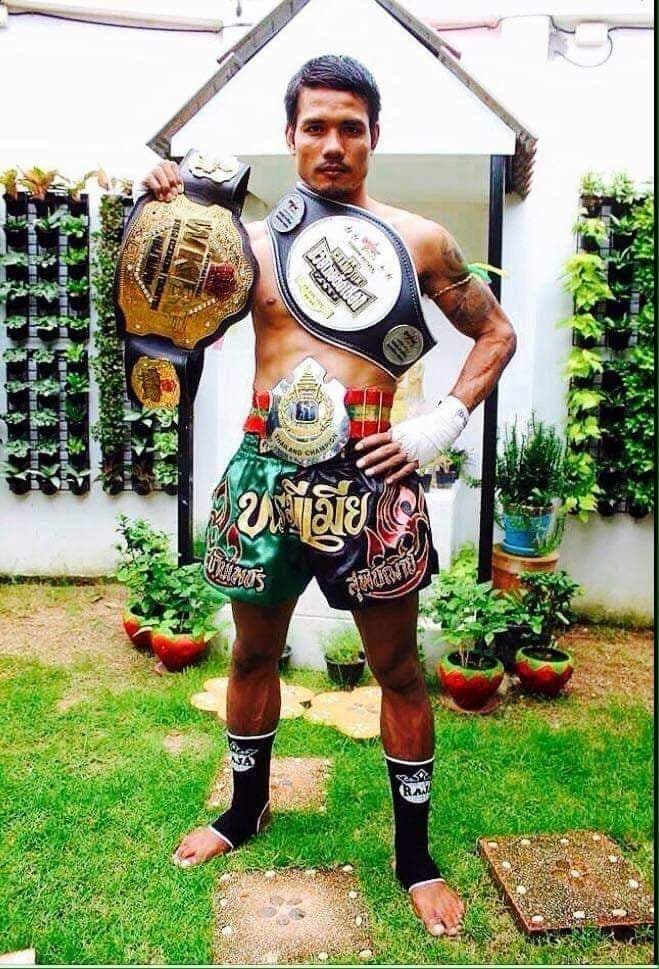 A male trainer in boxing attire poses with championship belts in an outdoor setting.