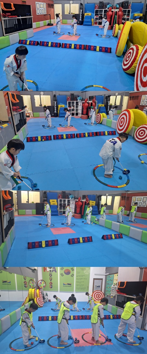 A brightly lit indoor space shows children in white uniforms practicing martial arts with a trainer, utilizing targets and mats.