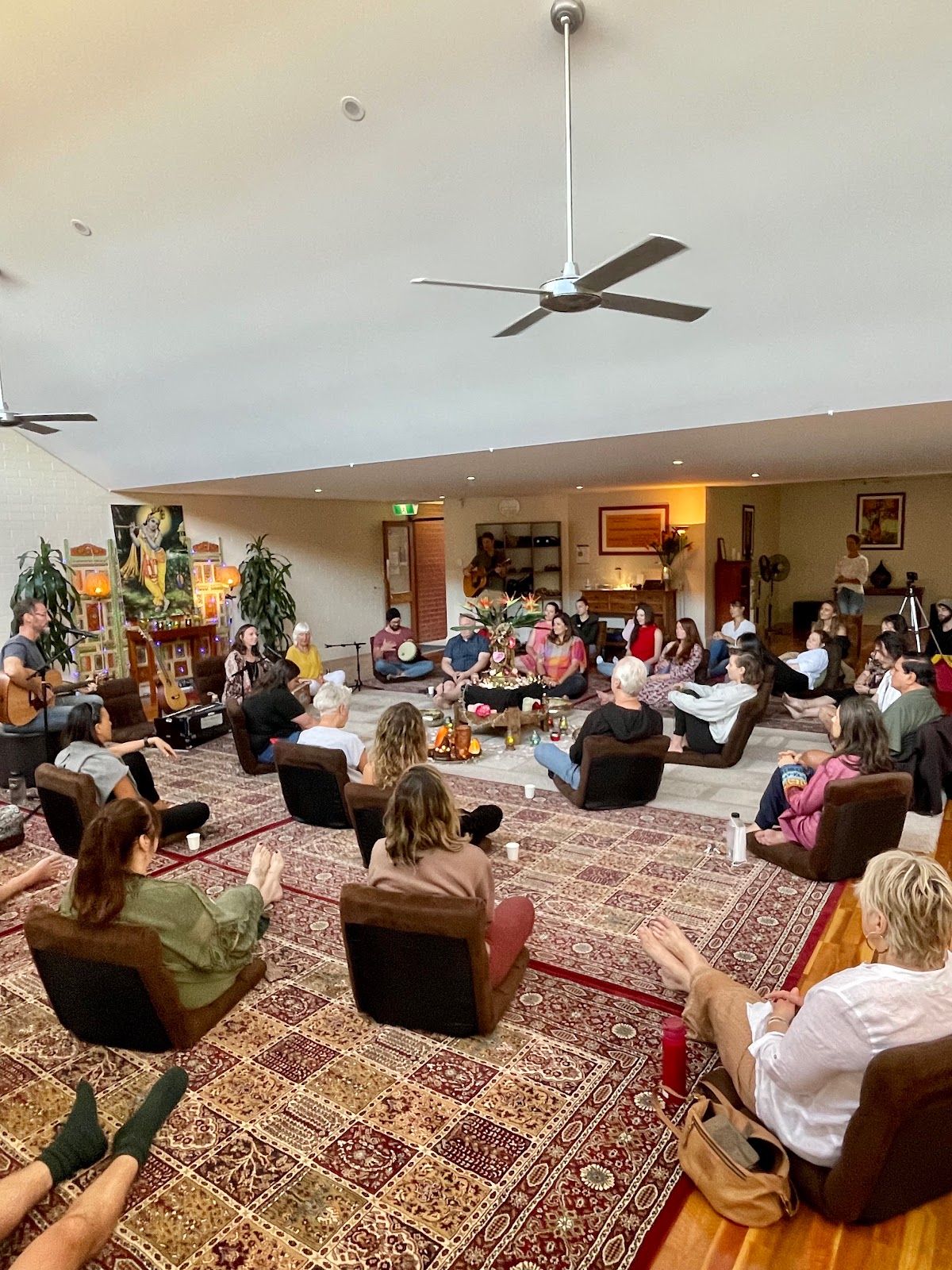 A group of people are seated in a circle on rugs and chairs, seemingly participating in a wellness or meditative activity in a warmly lit room.