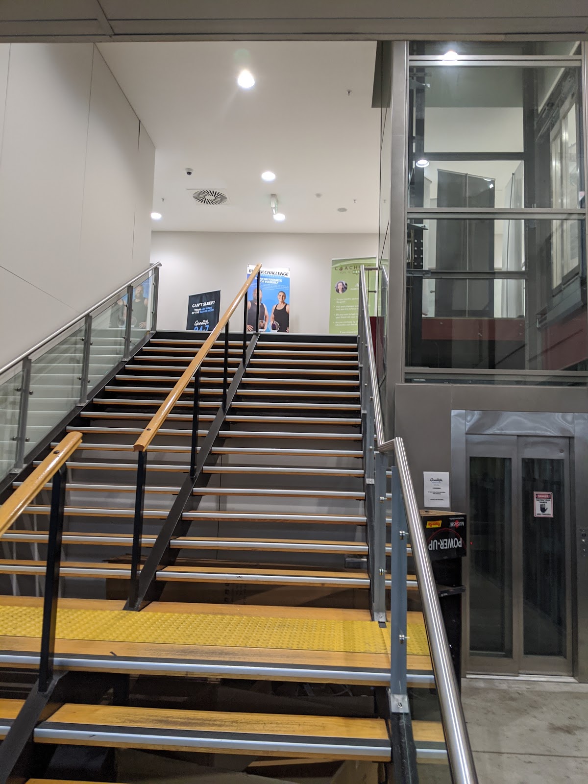 The photo shows a modern staircase leading up to what appears to be a fitness facility with glass doors and promotional posters visible.