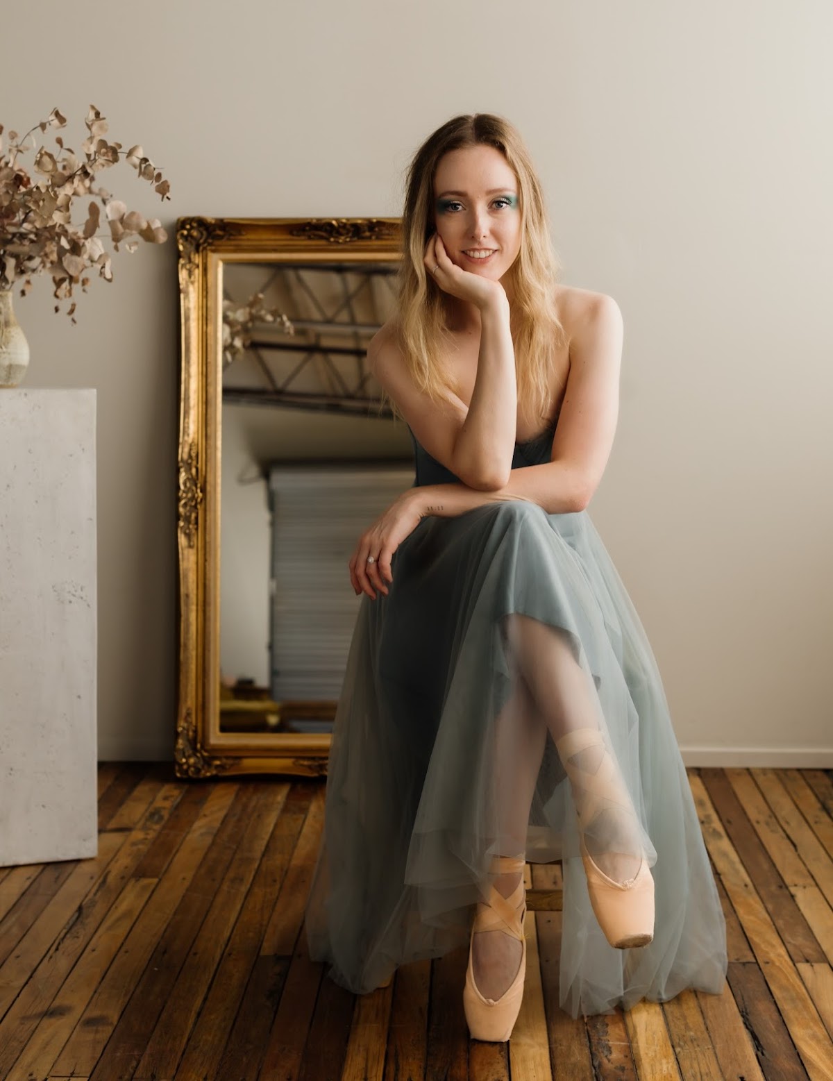 A portrait of a female trainer in ballet attire sitting on a wooden floor with a decorative mirror and concrete block backdrop.