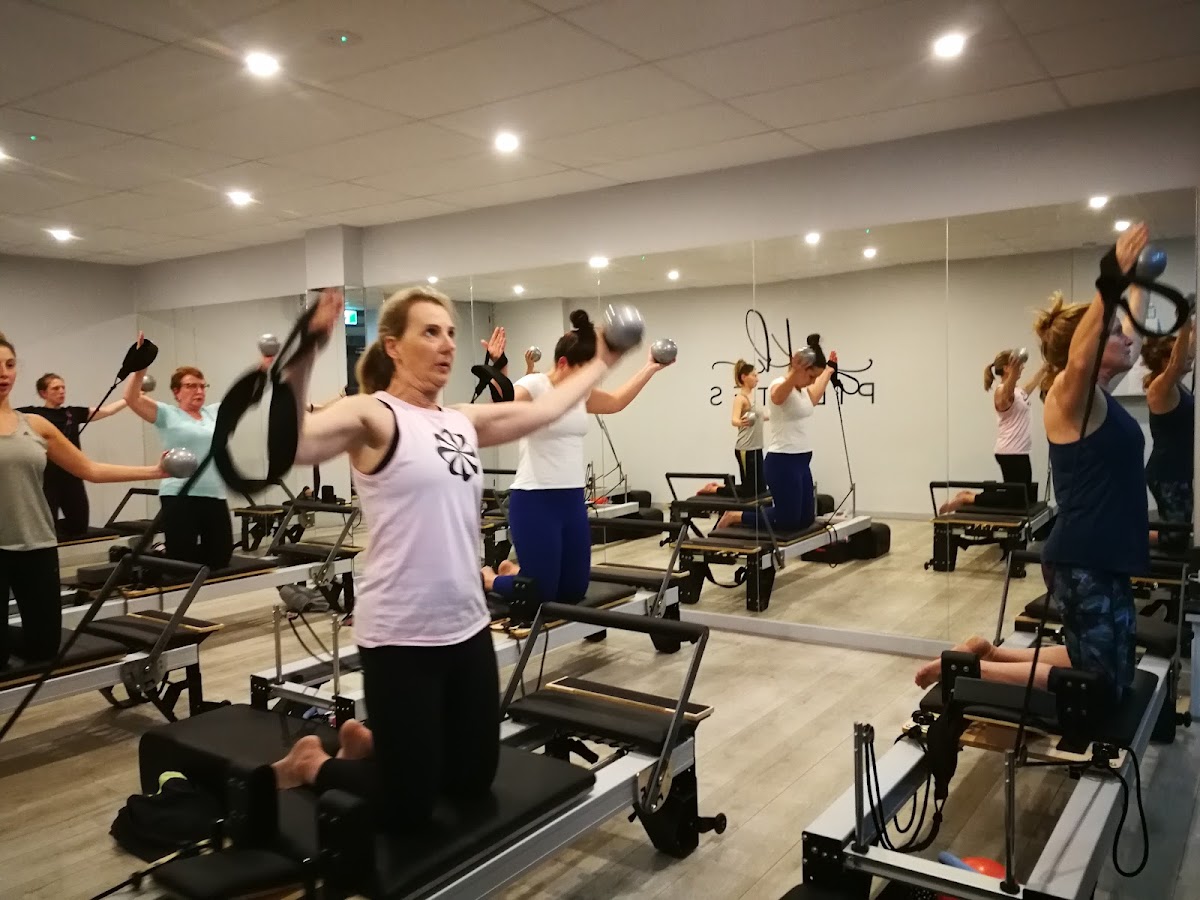 A group fitness class is in progress with participants using reformer pilates machines under the guidance of a female trainer.