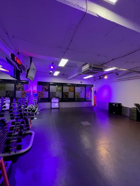 The photo shows a gym floor with free weights and industrial lighting in shades of purple.