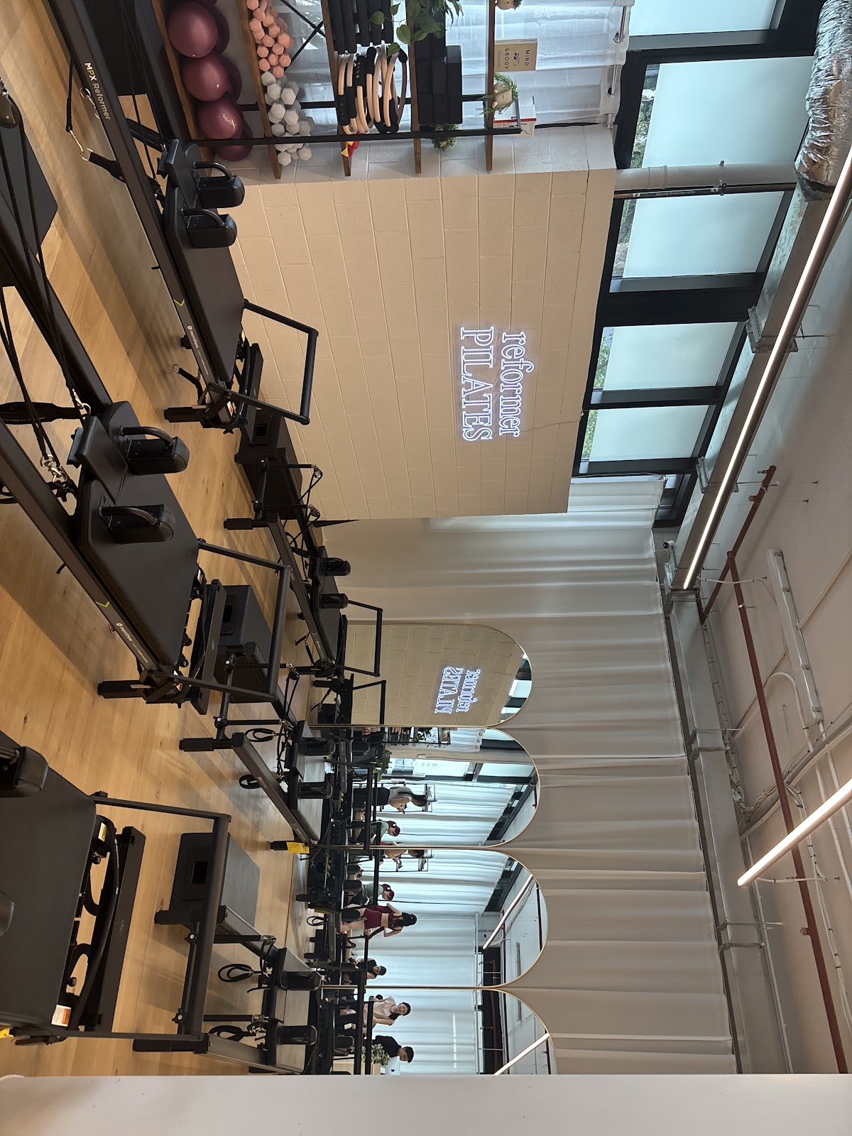 The photo shows an interior view of a fitness studio with several reformer pilates machines and people working out.