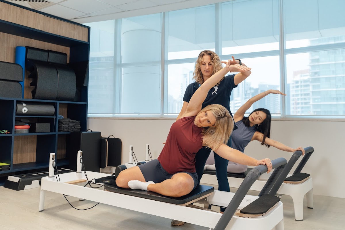 Three women are participating in a pilates session with a trainer using reformer machines in a bright, modern studio.