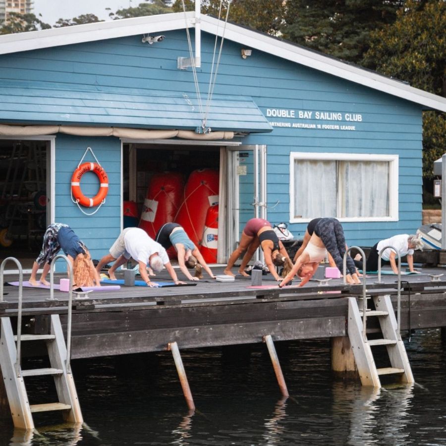 A group of people are practicing yoga on a wooden dock in front of a blue building with the words 'Double Bay Sailing Club' displayed.