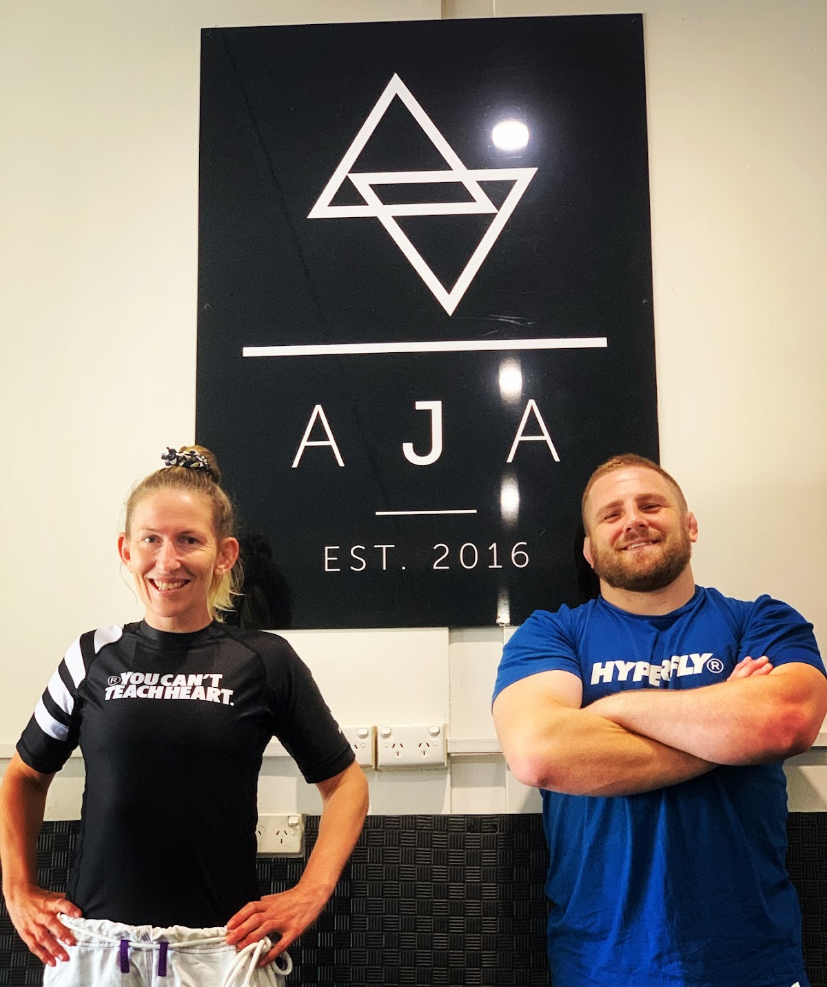 Two trainers, one male and one female, pose in front of a black sign with a logo and text, suggesting a fitness business.
