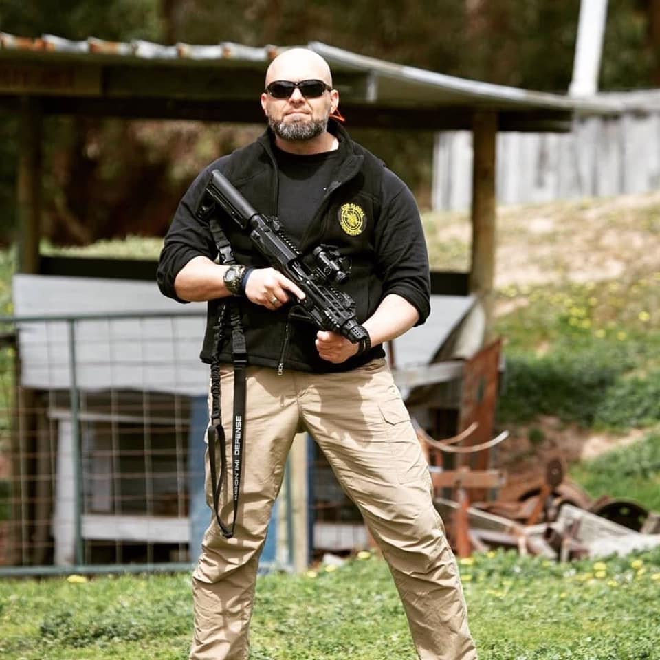 A portrait of a male trainer holding a rifle in an outdoor shooting range setting.