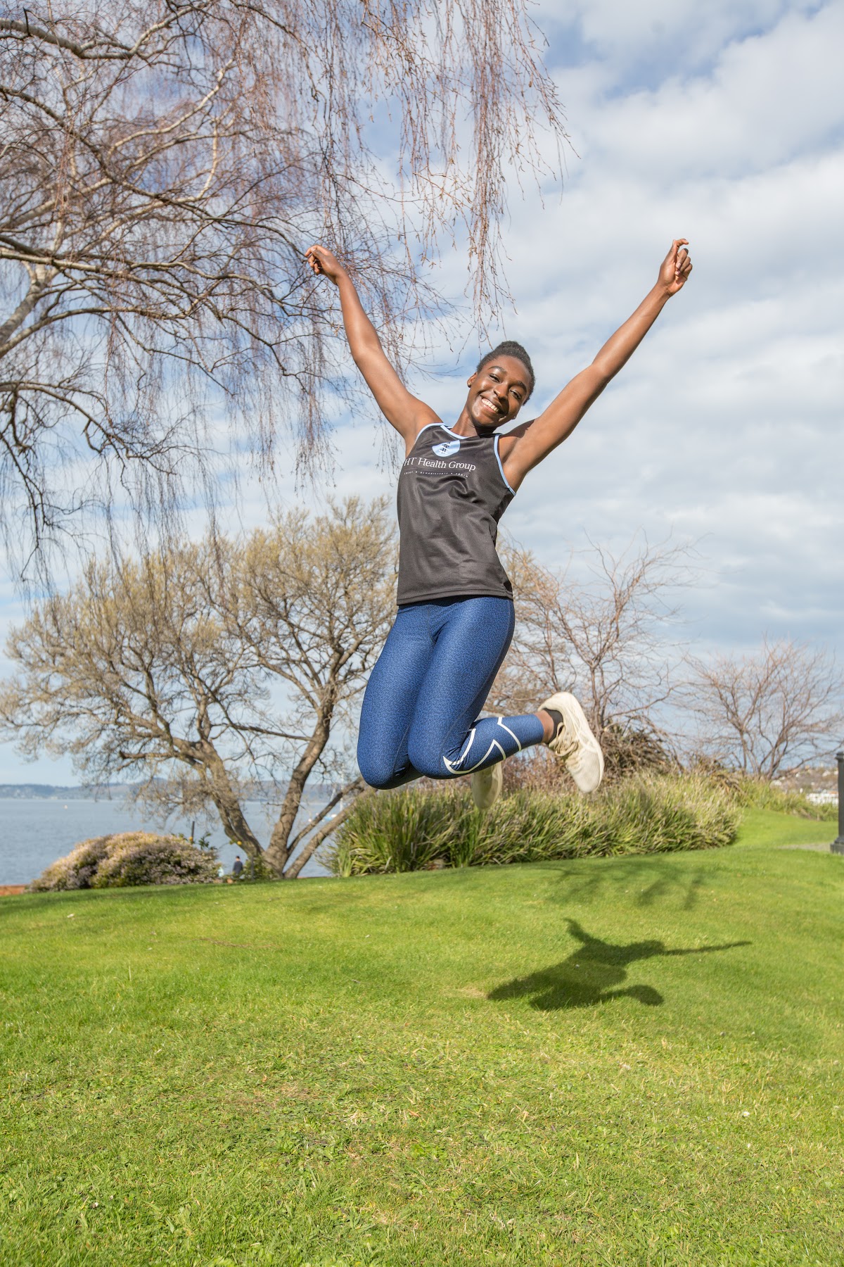 A woman jumping in mid-air over a grassy area near water, likely participating in an outdoor fitness activity.