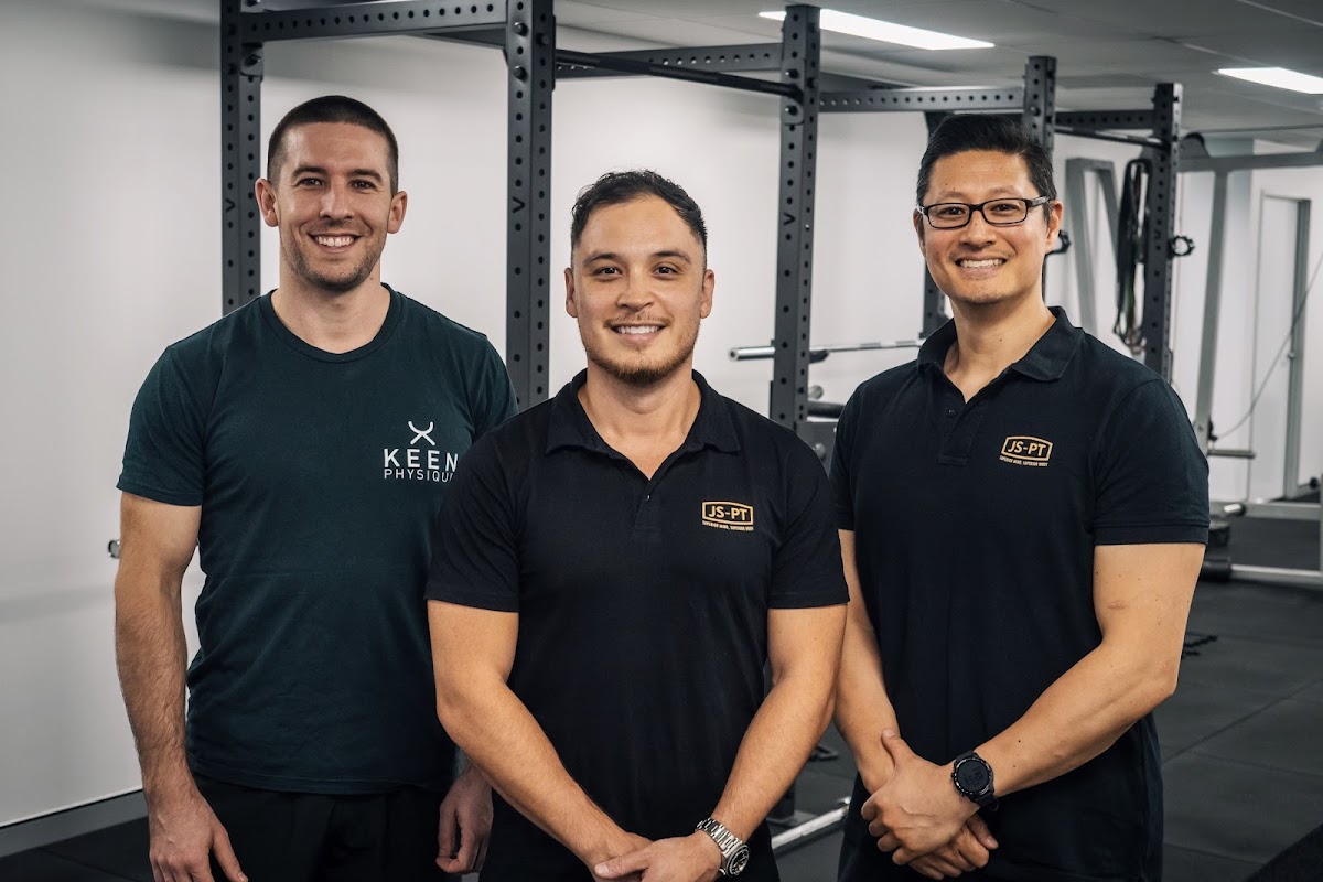 Three male trainers are posing in front of a squat rack within a clean and minimalist gym setting.