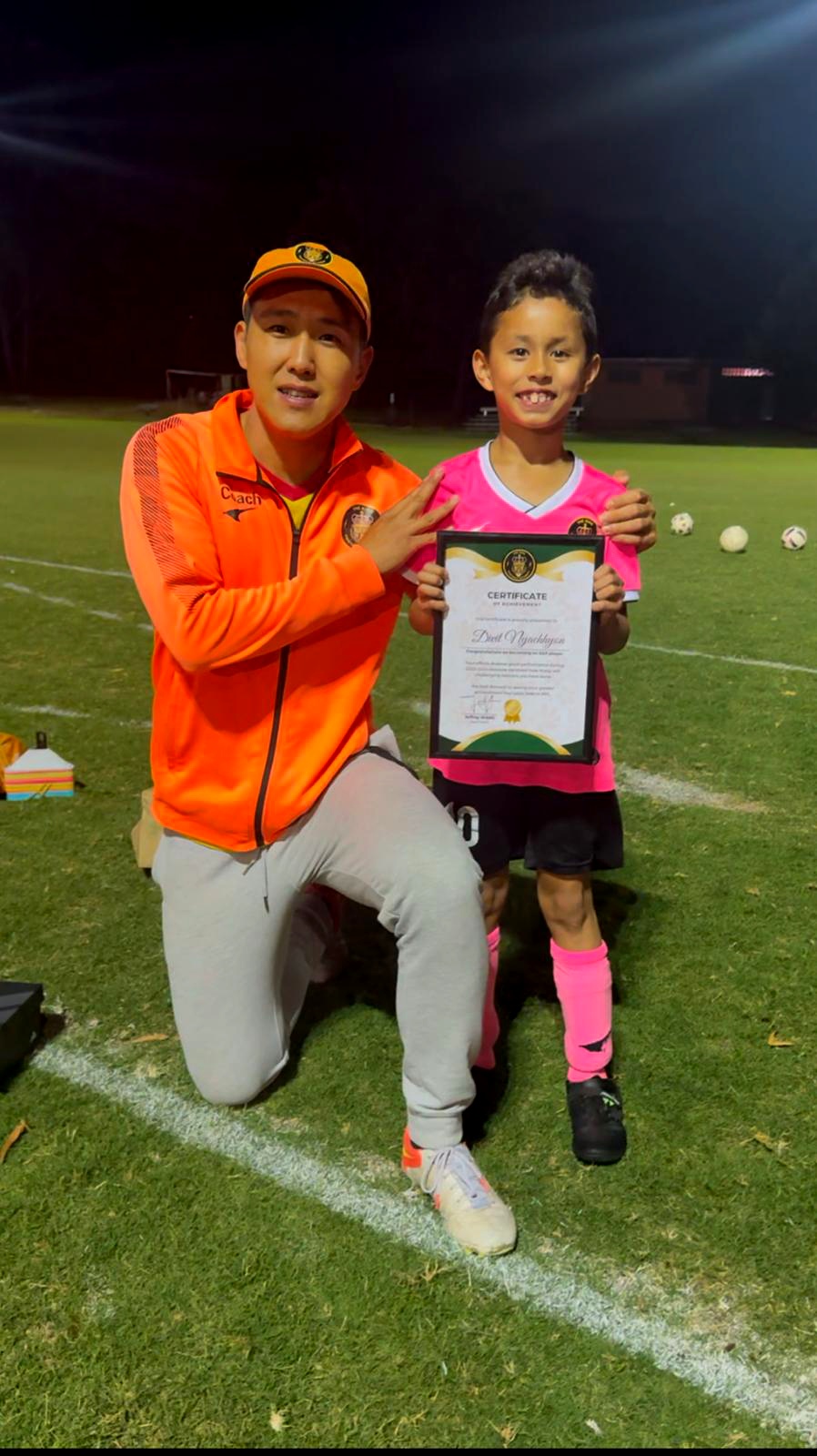 A male trainer is kneeling and posing with a young boy who holds a certificate, likely celebrating an achievement in martial arts or fitness.