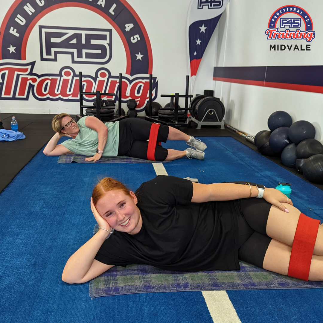 Two women are lying on the floor performing abdominal exercises in a brightly lit gym setting with free weights and a squat rack visible.