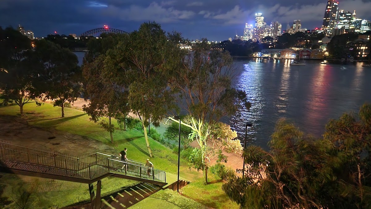 A nighttime view of an outdoor fitness area with people exercising on a staircase overlooking a river and city skyline.