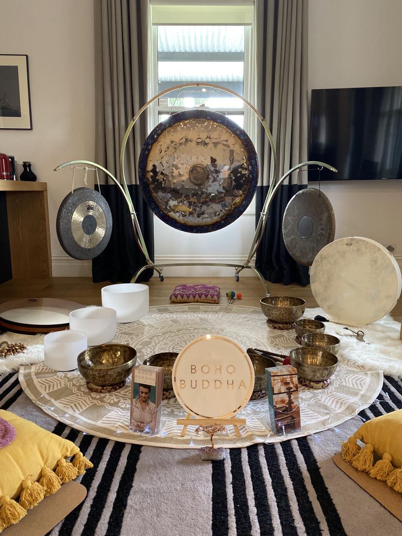 The photo depicts a serene studio space with yoga mats and sound healing instruments like gongs and singing bowls arranged for a meditative or therape