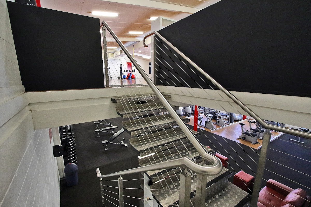 An elevated view of a gym floor with free weights and cardio machines visible below, framed by a staircase with metal railings.