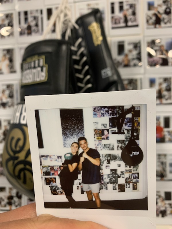 A close-up shot of boxing gloves hanging in a gym with a wall covered in photos, including a polaroid of two trainers.