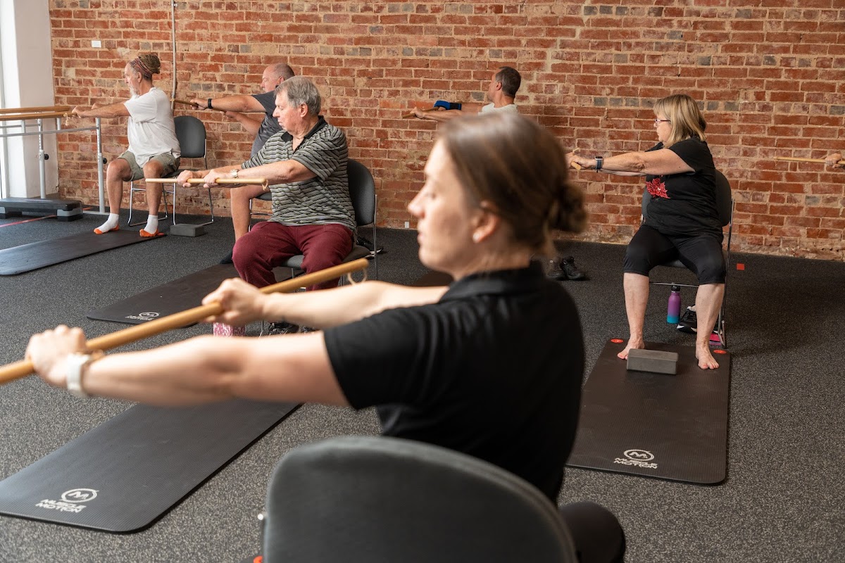A group of adults participates in a fitness class with a focus on strength and flexibility, utilizing yoga mats and light weights in a studio setting 