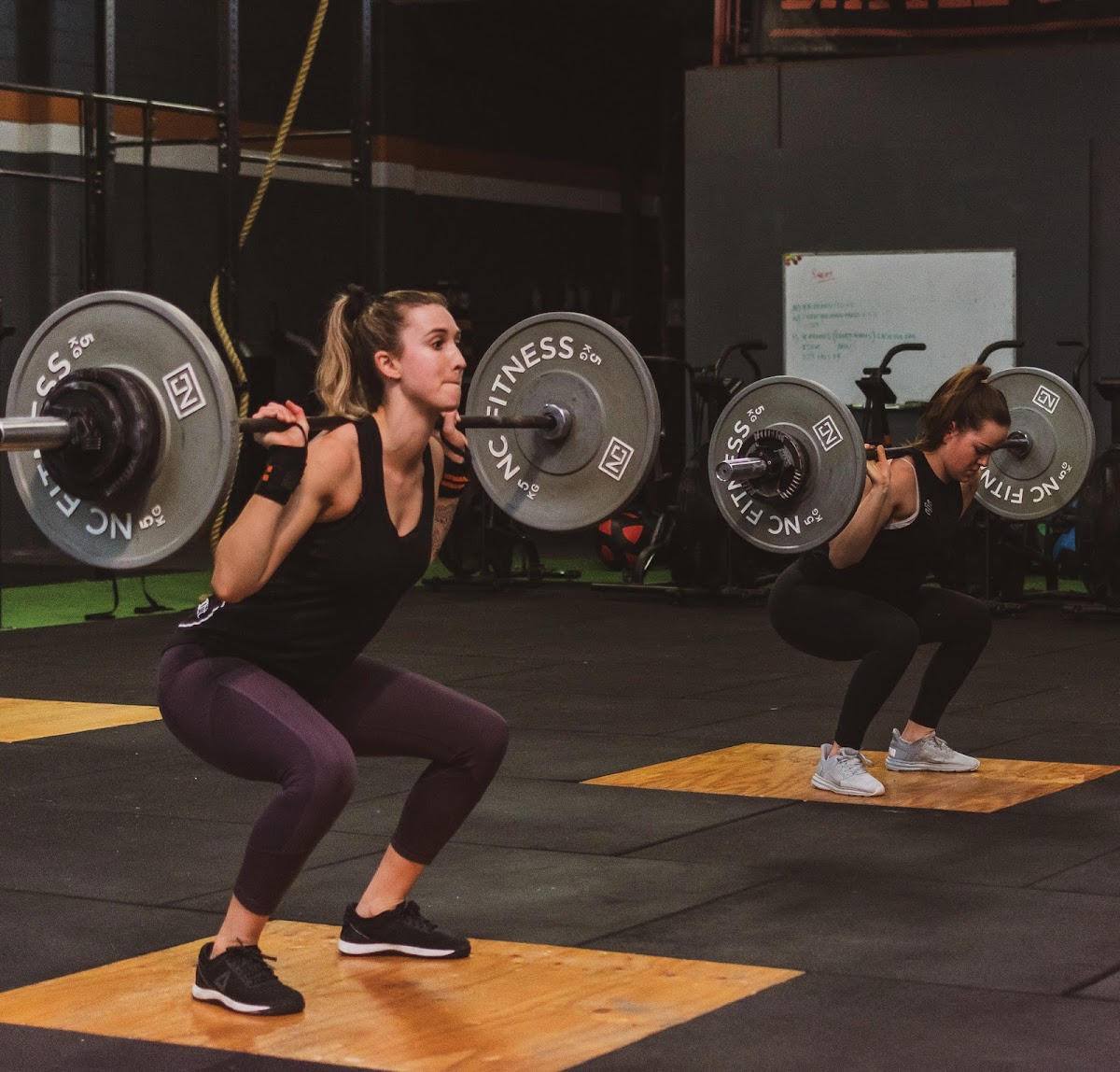 Two individuals are performing squats with barbells in a gym setting.