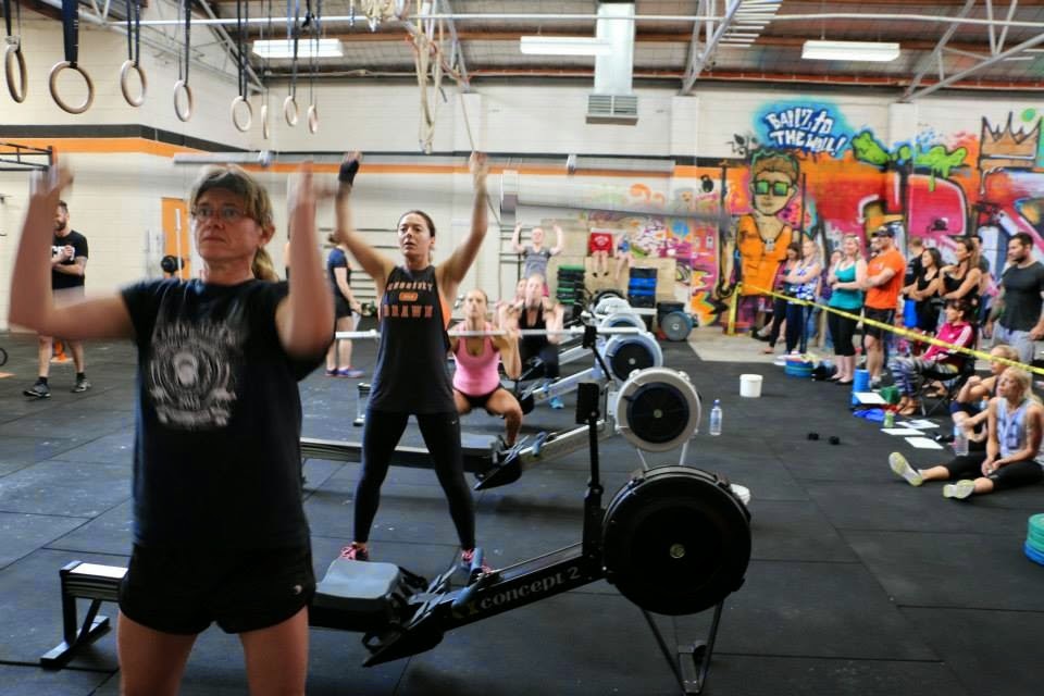 A group of people participate in a strength training class in an industrial-style gym, with a focus on rowing and weightlifting.