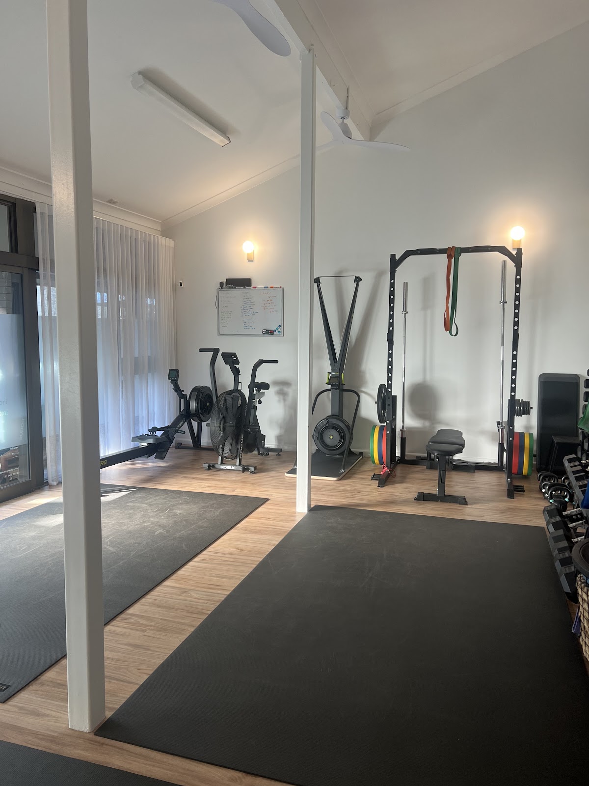 A minimalist gym space features a rowing machine, free weights, and TRX suspension trainer, with a clean and bright aesthetic.