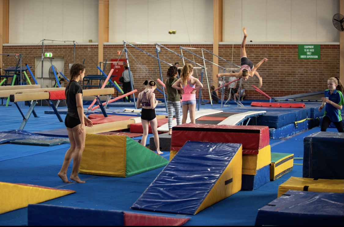 A group of young girls are participating in a gymnastics class, with a trainer supervising and guiding them through various exercises.