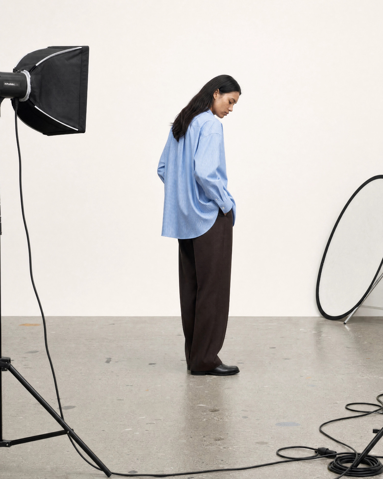 Fashion model in a loose blue striped shirt and wide brown trousers standing in a studio, surrounded by lighting gear and cables.