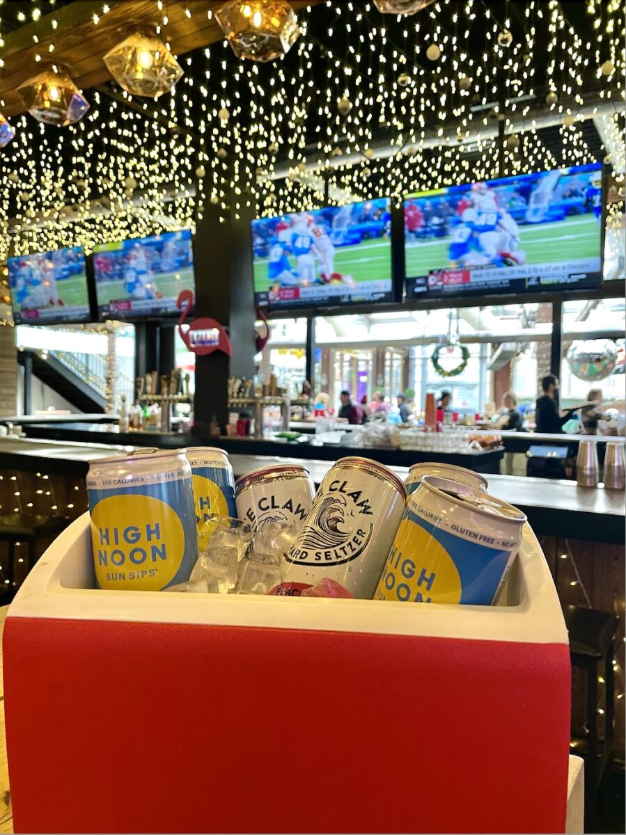 High Noons and White Claws in a red cooler with football playing on the screens at the bar in the background.