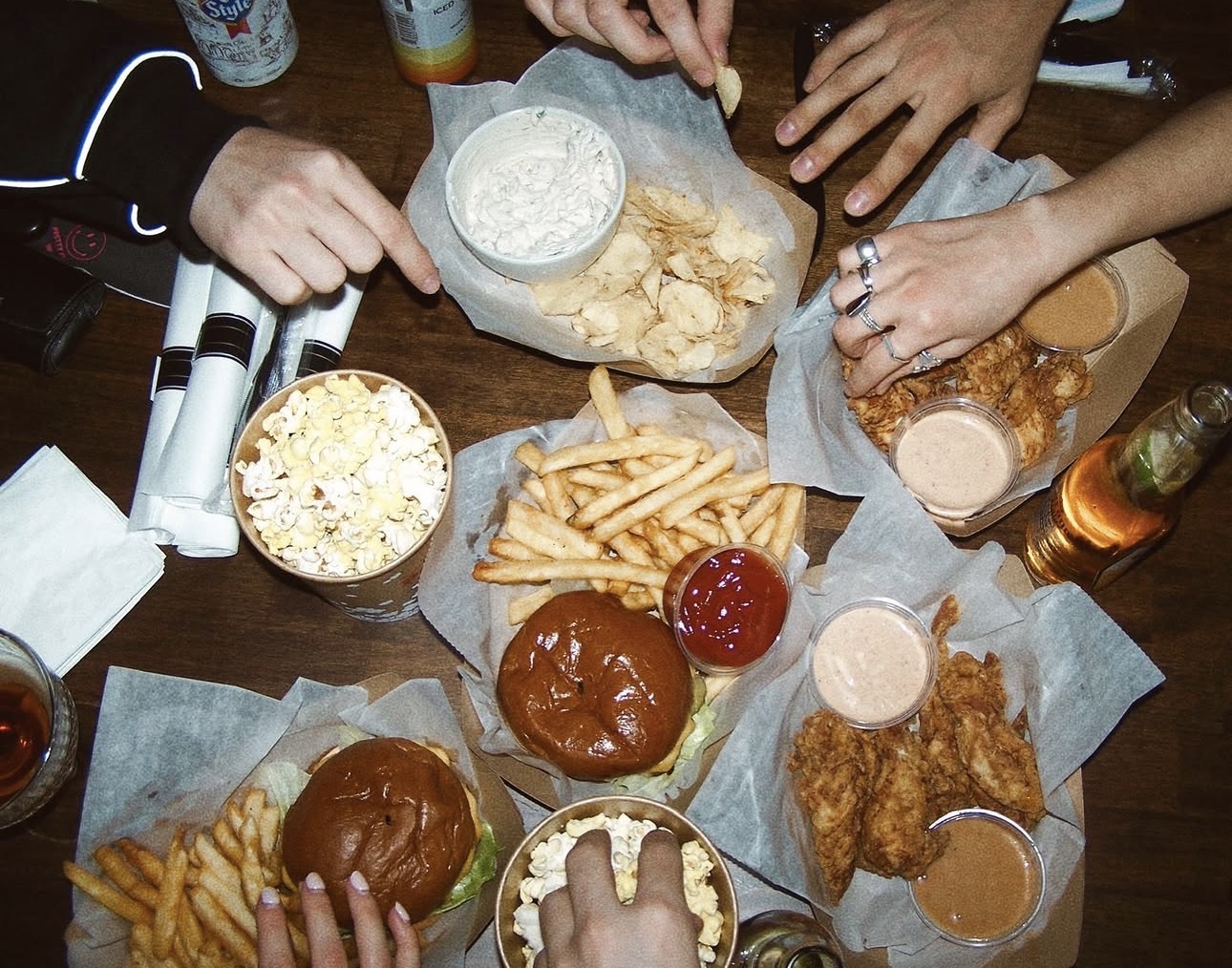 Table spread of bar food including burgers, fries, fried chicken tenders, chips with dip, popcorn, and drinks shared by a group.