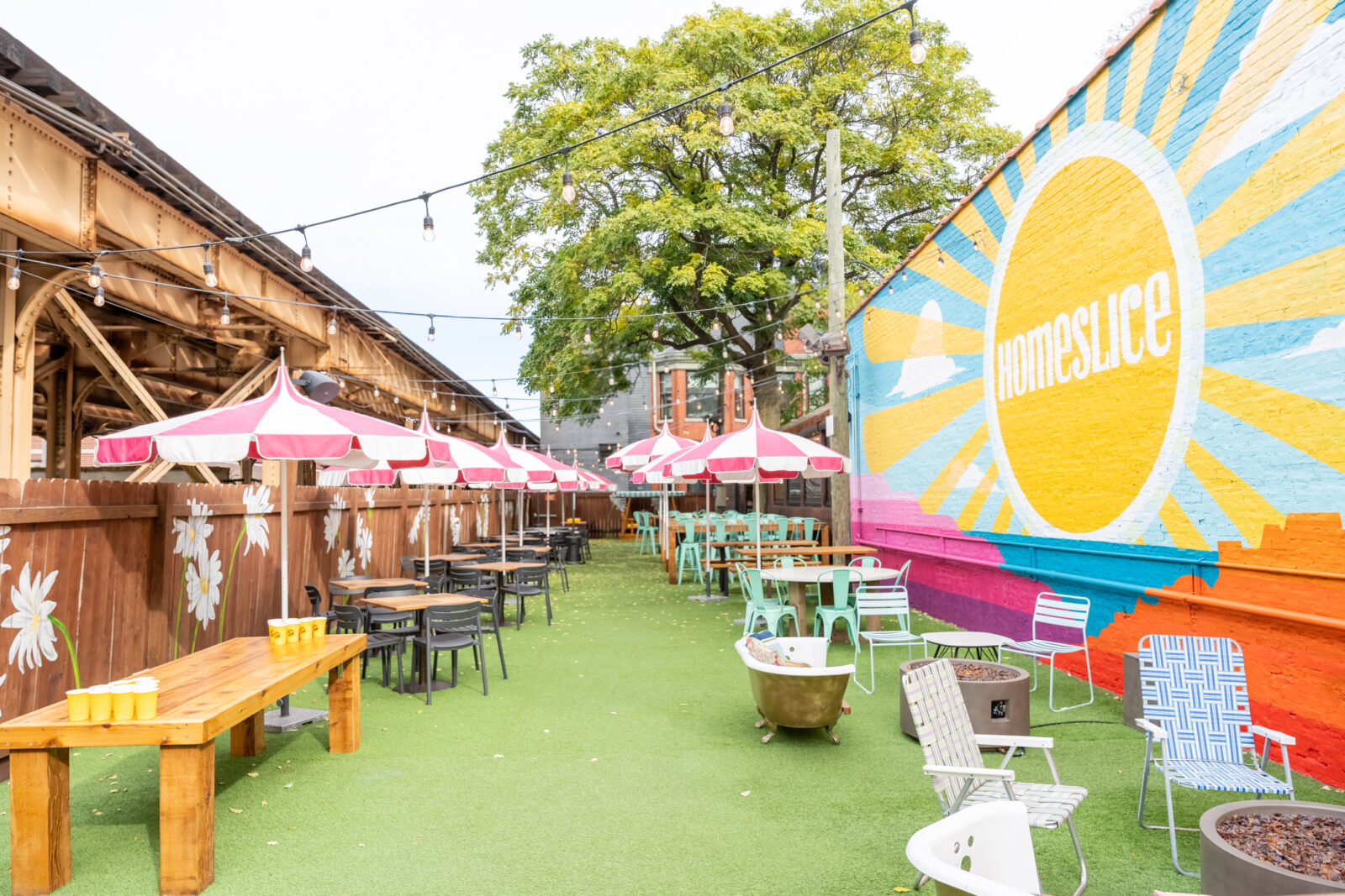 slide 4 of 4 - Outdoor patio, pink and white umbrellas over high and low top tables, large homeslice sun mural, beer pong