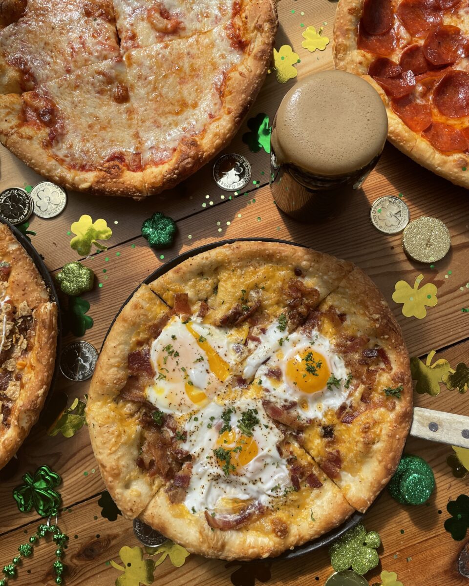 A festive St. Patrick’s Day spread featuring multiple pizzas, including a breakfast pizza topped with eggs, bacon, and cheese. A pint of dark beer sits among gold coins, green shamrocks, and glittery decorations on a wooden table, creating a celebratory Irish-inspired scene.