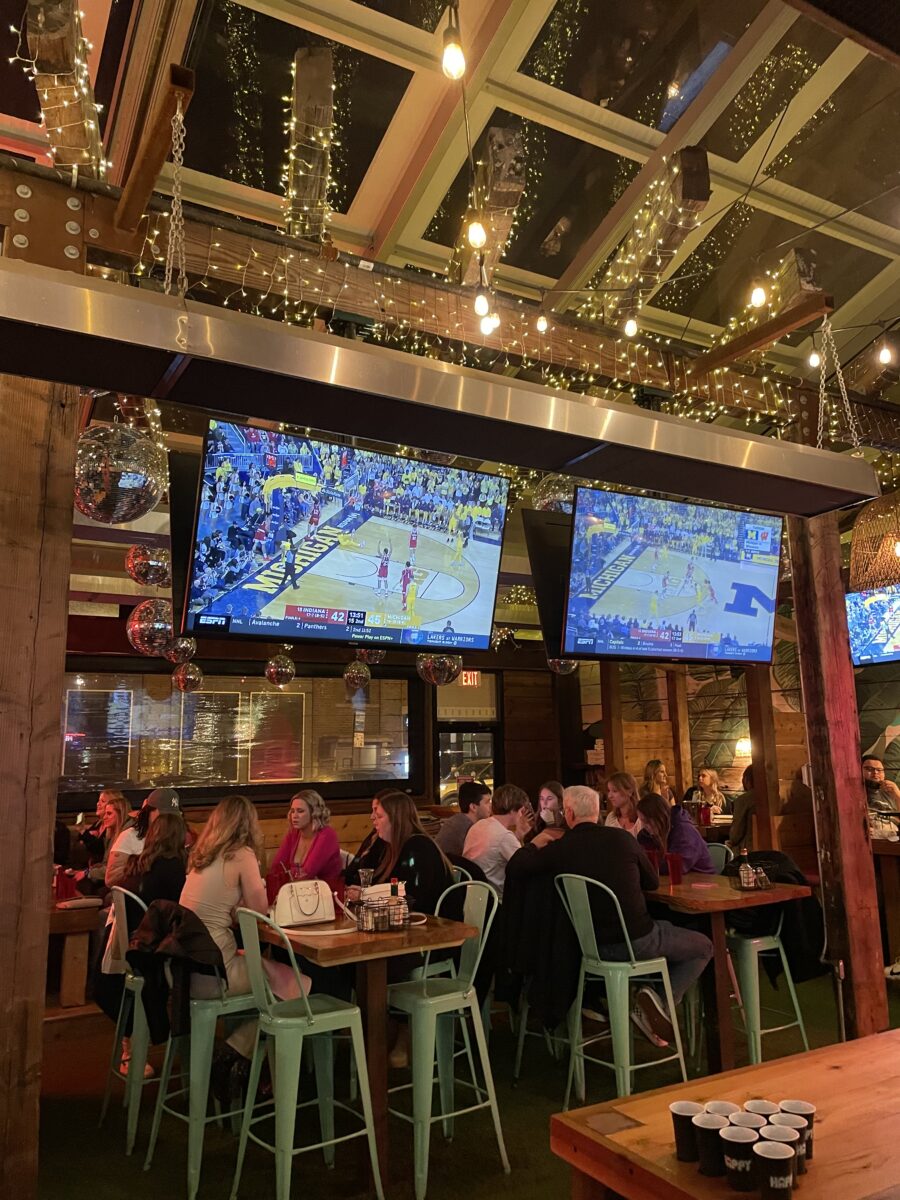 Busy bar interior with groups of people seated at tables watching basketball on large TVs, string lights hanging from the ceiling, and a lively, social atmosphere.