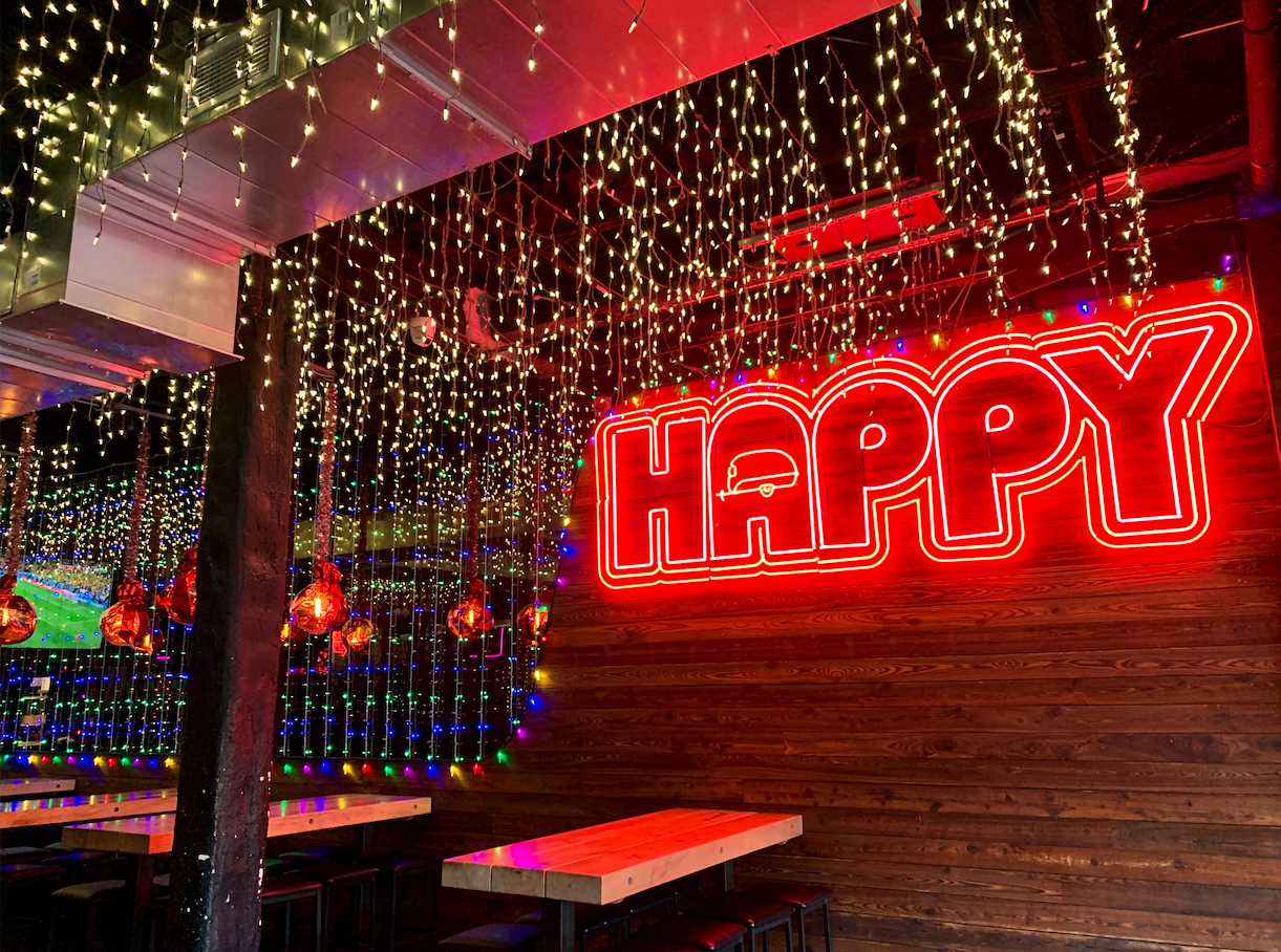 happy camper logo neon sign hanging above seating area with Christmas lights hanging from the ceiling and TVs playing sports