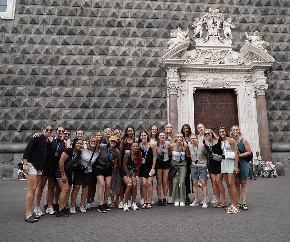 Student athletes and coaches of the Iowa Women's Basketball team stand together in front of the Gesù Nuovo in Naples, Italy