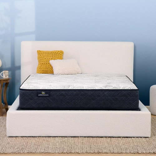 A straight-on view of a Serta Perfect Sleeper mattress featuring a white top with a grey fan-like pattern and dark navy sides. The bed is styled with a yellow knit pillow and a small white textured pillow against an off-white boucle headboard.