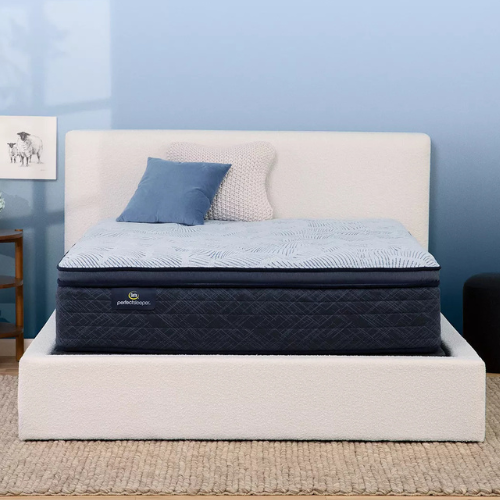 Close-up of a Serta Perfect Sleeper mattress featuring a dark navy quilted side with a prominent brand label. The top surface displays a decorative blue sunburst pattern. It is set within a textured off-white upholstered bed frame and styled with decorative pillows against a light blue wall.