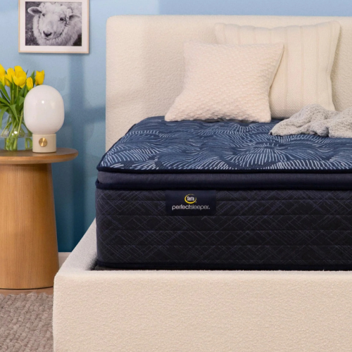 A close-up photograph shows a Serta Perfect Sleeper mattress with a dark blue patterned top and navy quilted sides, featuring a prominent brand label. It is set within a textured, off-white upholstered bed frame and propped with two cream-colored decorative pillows against a light blue wall. To the left, a wooden side table holds a globe lamp and yellow tulips.